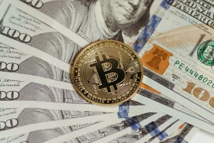 Moeda de Bitcoin sobre uma pilha de cédulas de cem dólares, representando a relação entre ativos digitais e financeiros tradicionais.