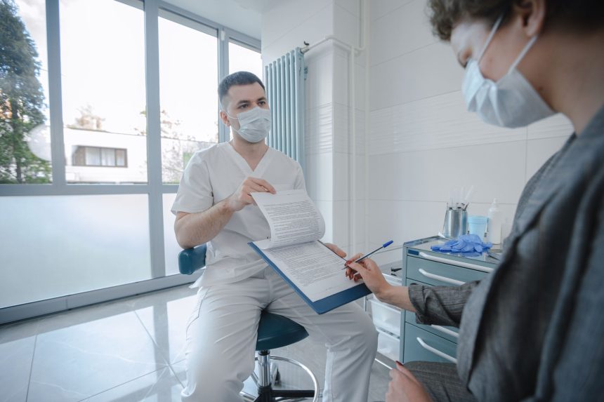 Médico e paciente conversam sobre um documento em uma clínica, ambos usando máscaras.